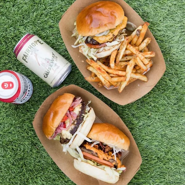 Two Ox Kitchen burgers and fries in paper trays, with two cans of Fairhope beer on green grass.