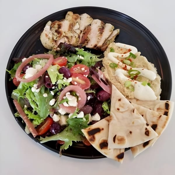 Grilled chicken, Greek salad, hummus, and pita bread on a black plate.