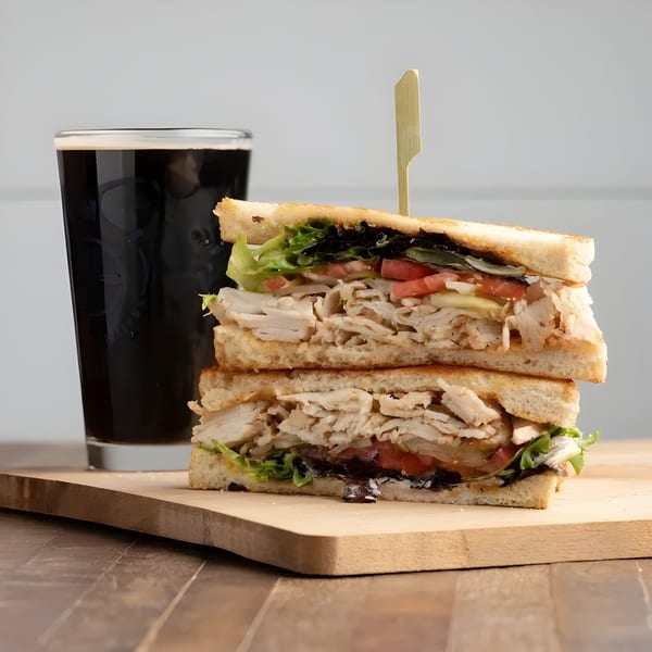 A stacked turkey club sandwich with lettuce and tomato, next to a dark beer on a wooden board.