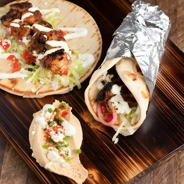 Two chicken pita wraps, one open and one foil-wrapped, with a side of hummus on a wooden board.