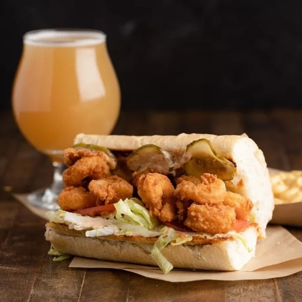 A fried shrimp po'boy with pickles, lettuce, and tomato, next to a glass of beer.