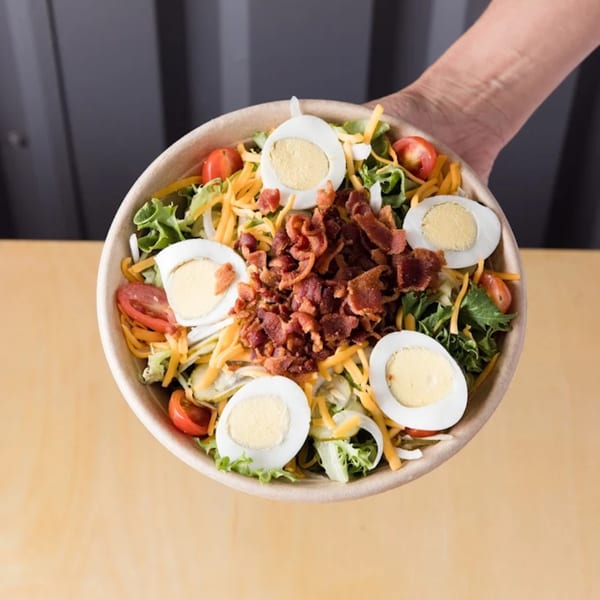 A hand holds a bowl of Ox Kitchen's Cobb salad with lettuce, bacon, eggs, cheese, and tomatoes.