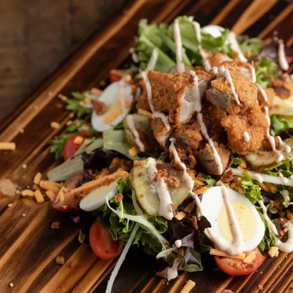 Ox Kitchen's fried chicken salad with lettuce, tomatoes, eggs, pickles, and dressing on a wooden board.